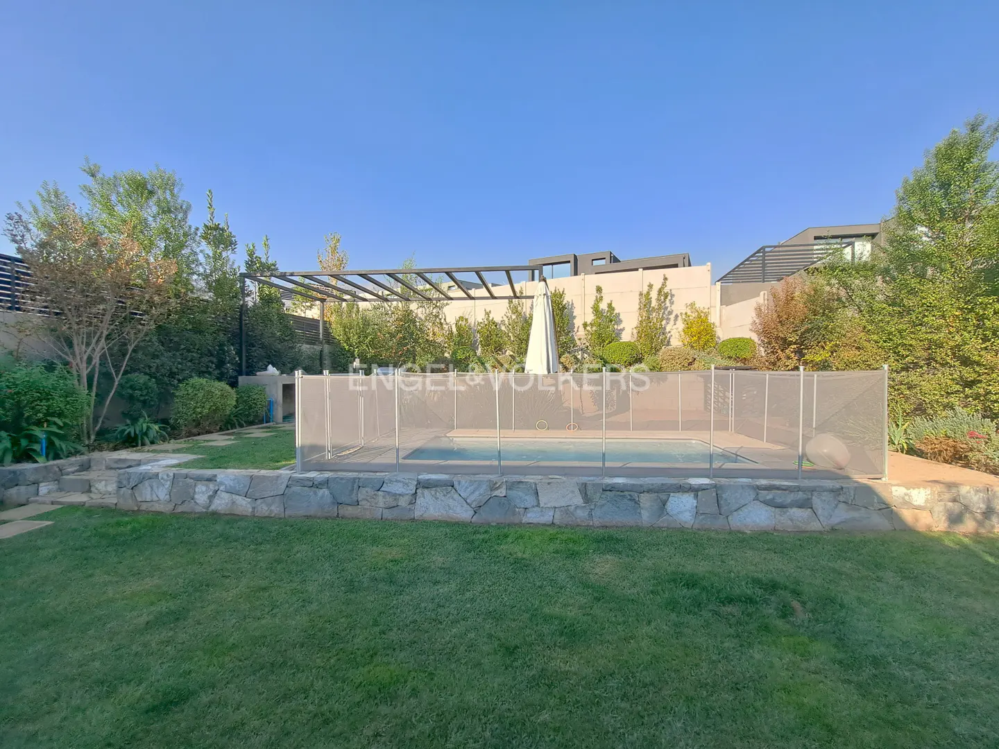 A backyard with a pool, enclosed by a mesh fence, sits on a stone base. Green grass covers the foreground, with trees and a pergola in the background under a clear blue sky.