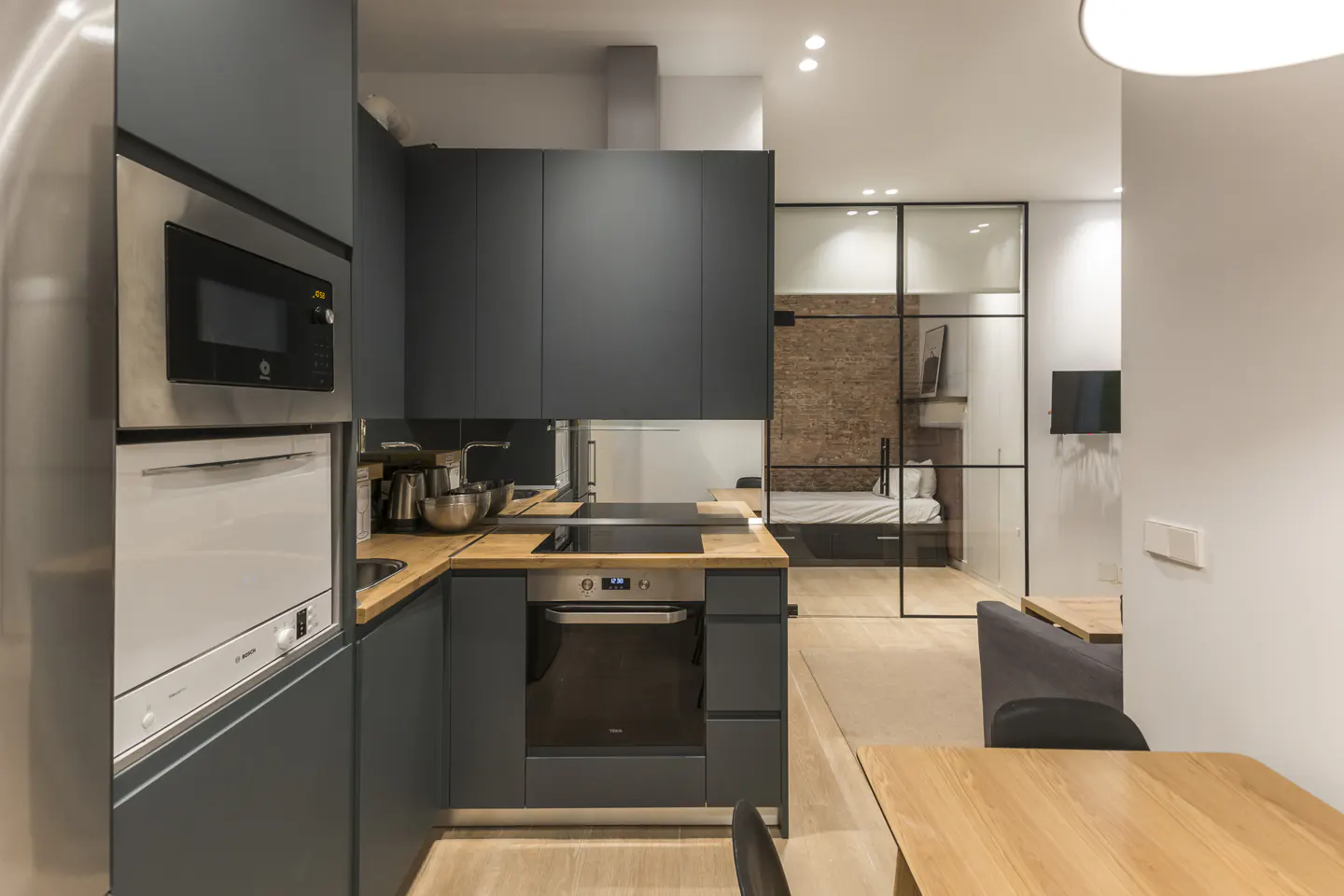 A modern apartment interior featuring a gray kitchen with wood countertops, stainless steel appliances, and a glass-enclosed bedroom.