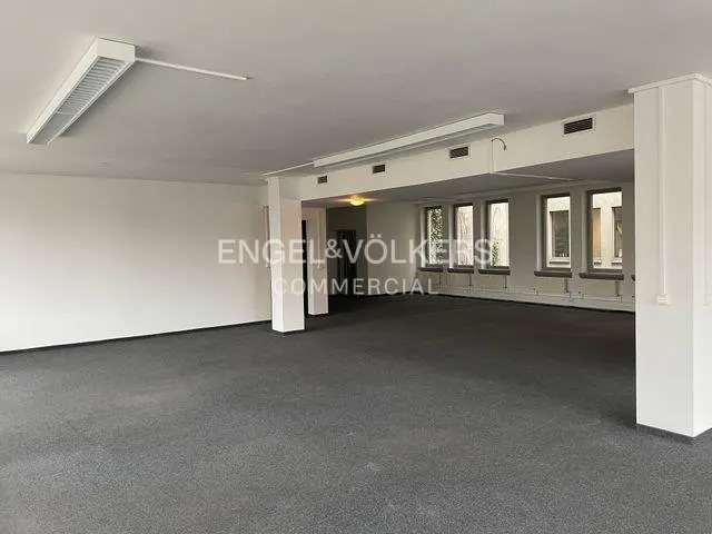 Empty, spacious commercial office with white walls, grey carpet, and large windows along the side wall.