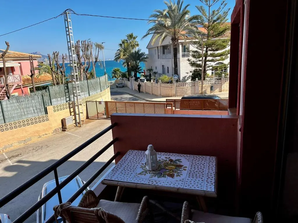 Balcony view of a street leading to the ocean. A table with a floral tile top and a lantern sits on the balcony. Palm trees and buildings are visible.