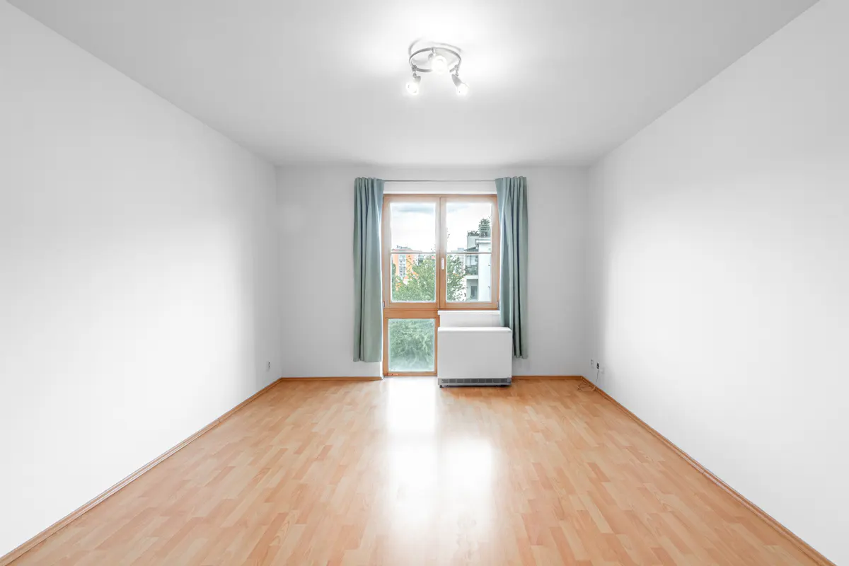 Empty room with white walls, light wood flooring, a window with blue curtains, and a white radiator.
