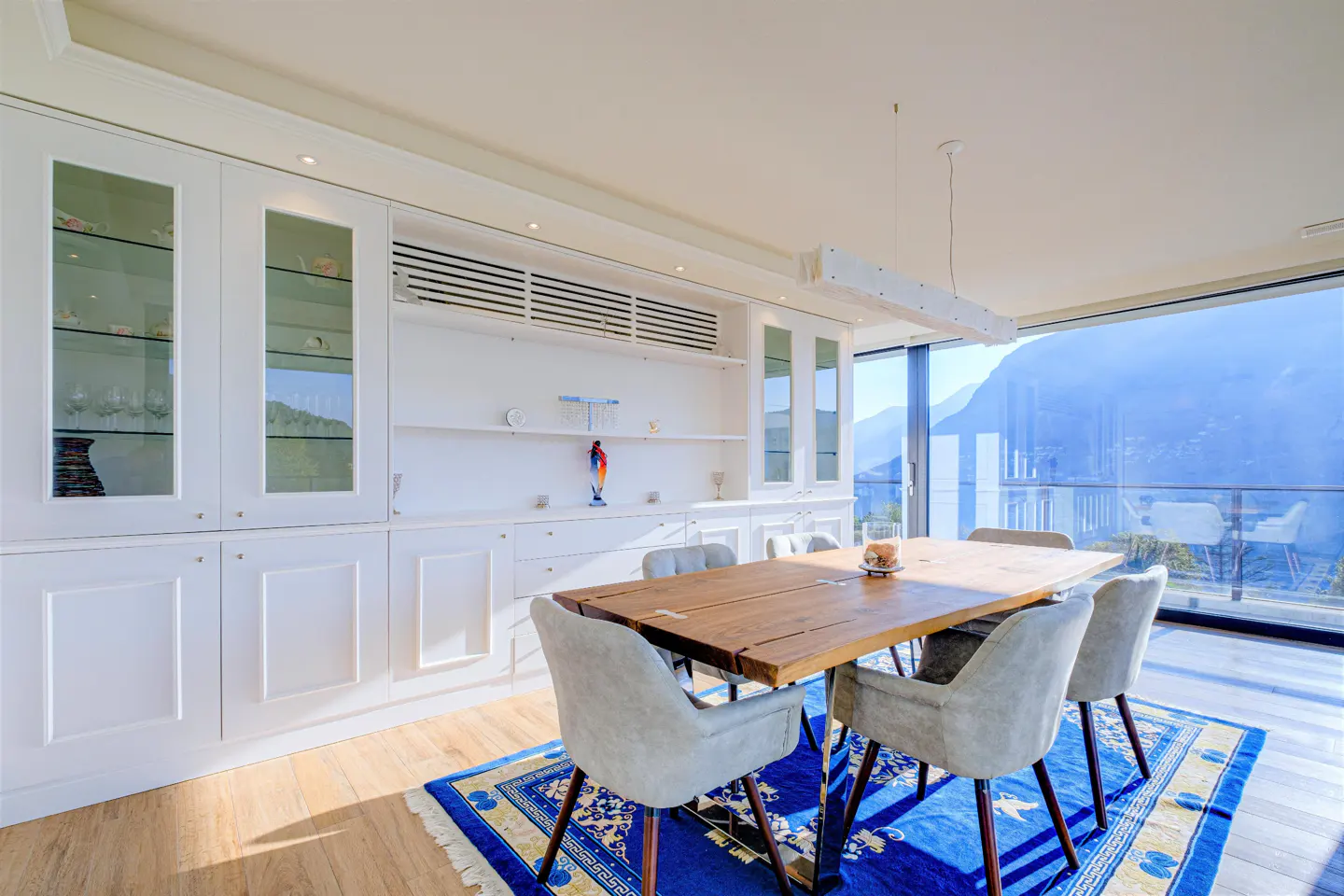 Bright dining room with a wood table, gray chairs, and a blue rug. White cabinets line the wall, and a large window shows a mountain view.