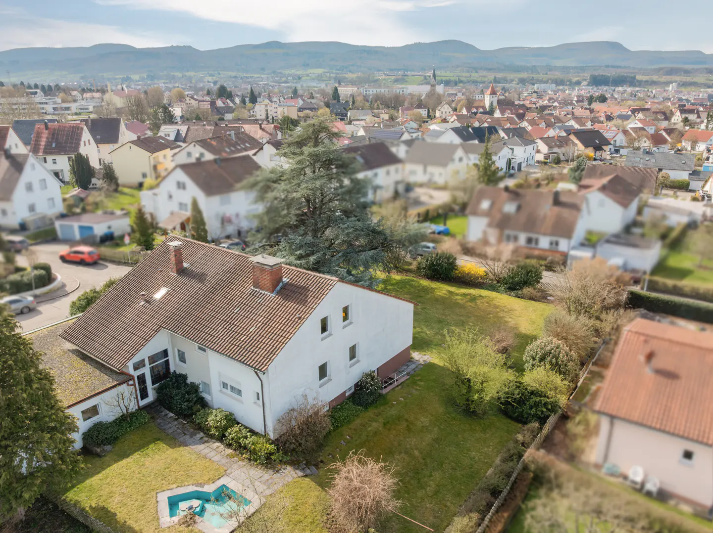 Aerial view of a white house with a brown roof, green lawn, and a small pool in the backyard. A town and mountains are in the background.