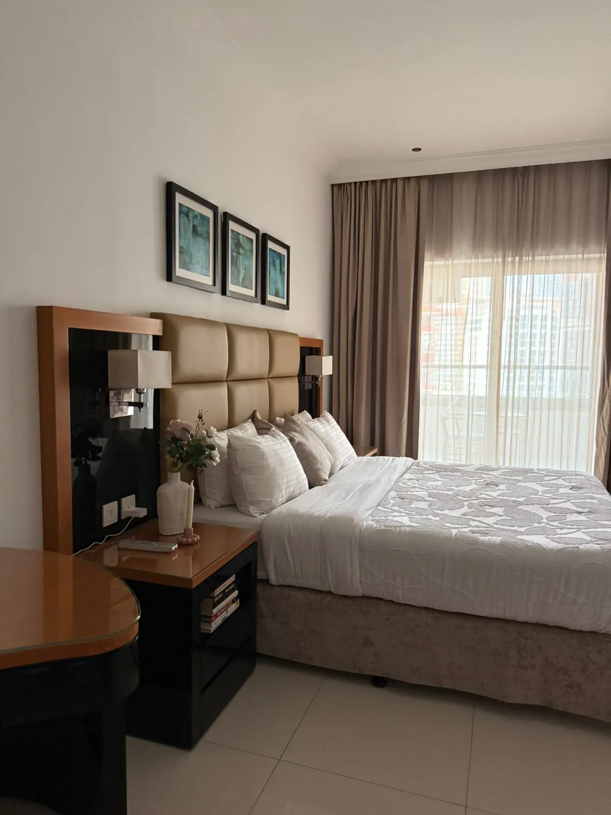 A bedroom with a large bed, tan headboard, white bedding, and a black nightstand with a lamp and flowers. Three framed pictures hang above the headboard. Curtains cover a window.