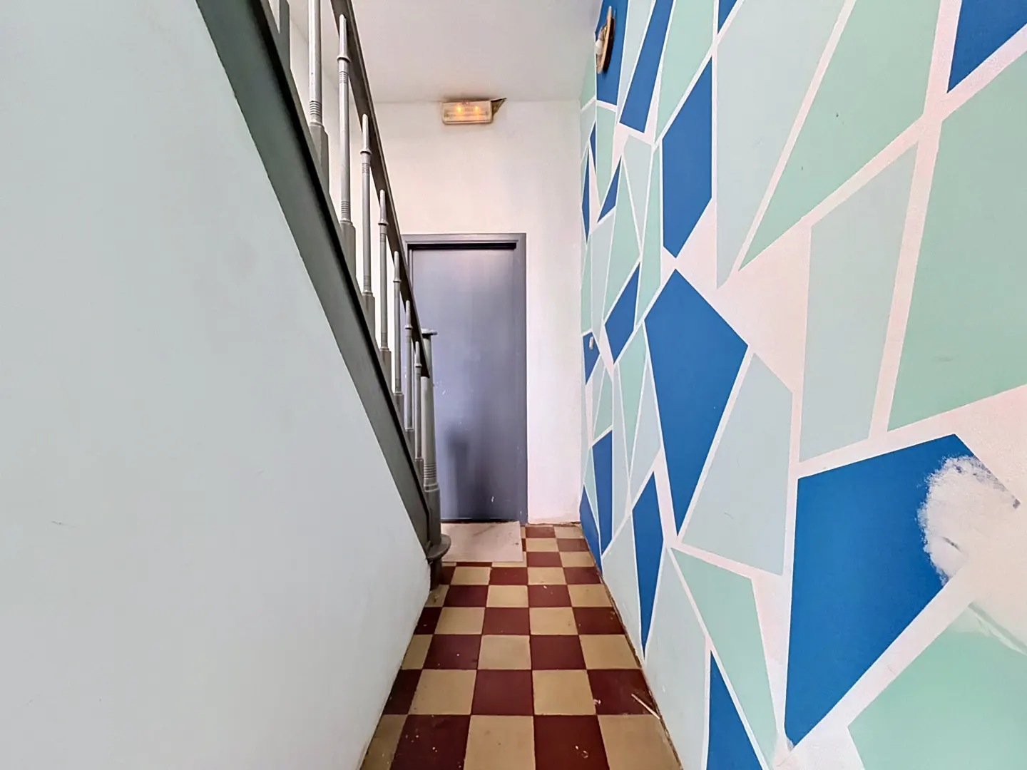 Hallway with stairs, a gray door, and a wall with blue and light green geometric shapes. The floor has a red and beige checkerboard pattern.