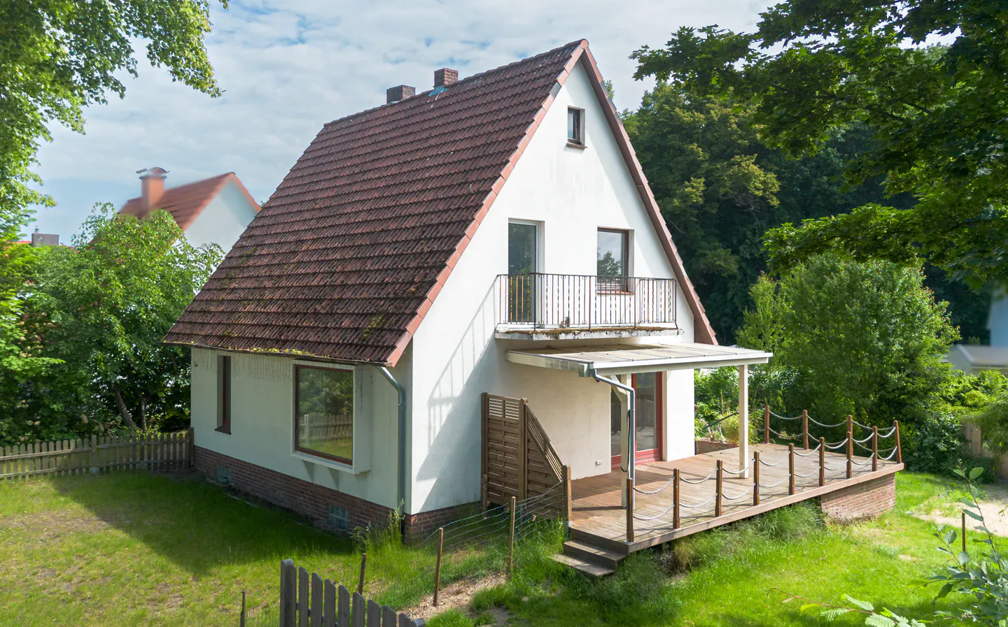 White house with a brown tiled roof, a small balcony, and a wooden deck in a lush, green backyard setting.
