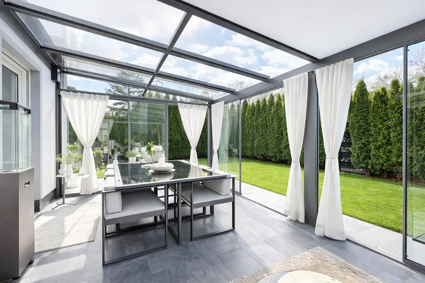 Sunroom with glass ceiling and walls, white curtains, black table and chairs, and green lawn outside.