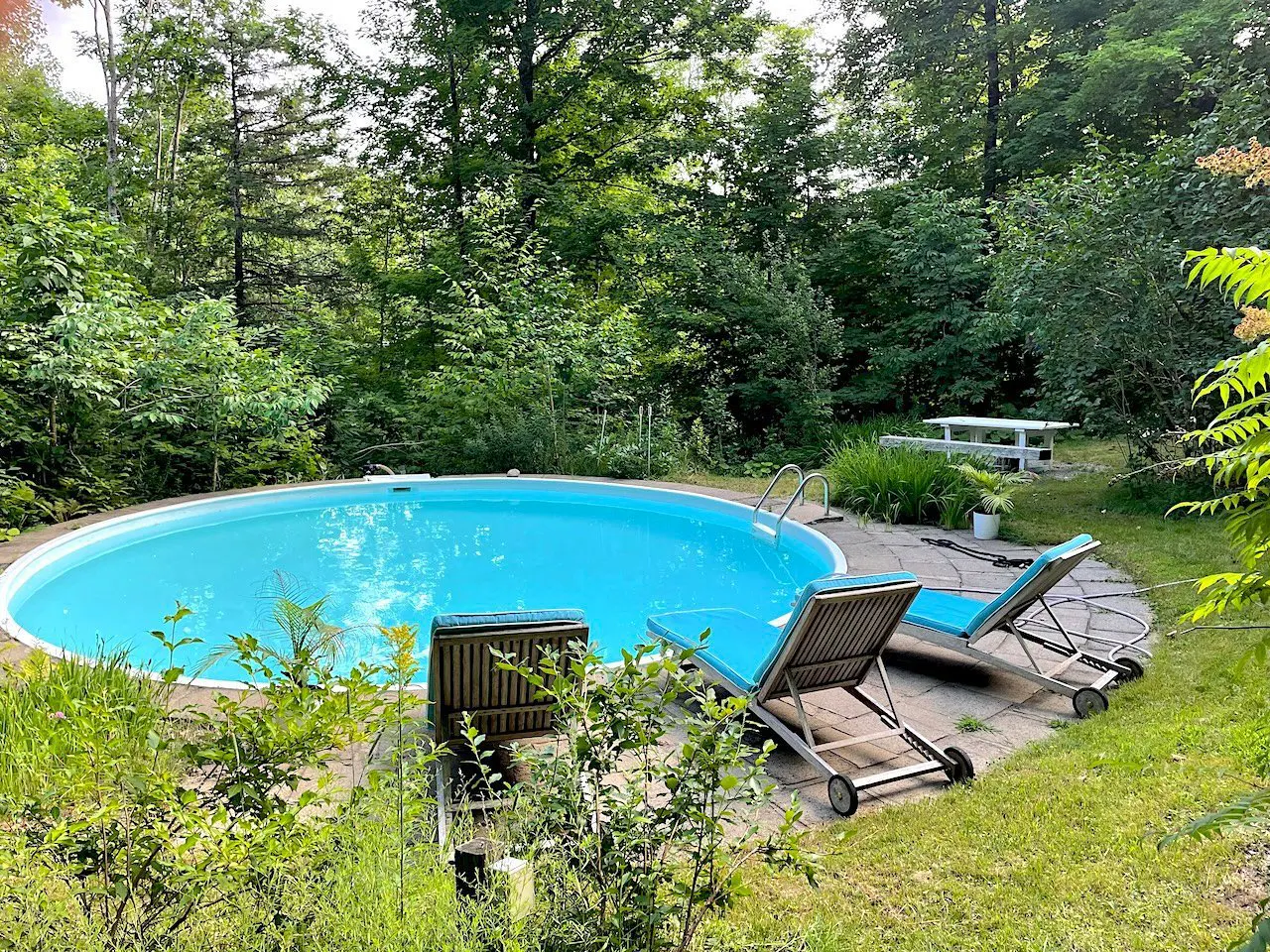 Outdoor pool with turquoise water, surrounded by lush greenery. Two lounge chairs with blue cushions sit on a stone patio. A white picnic table is visible in the background.
