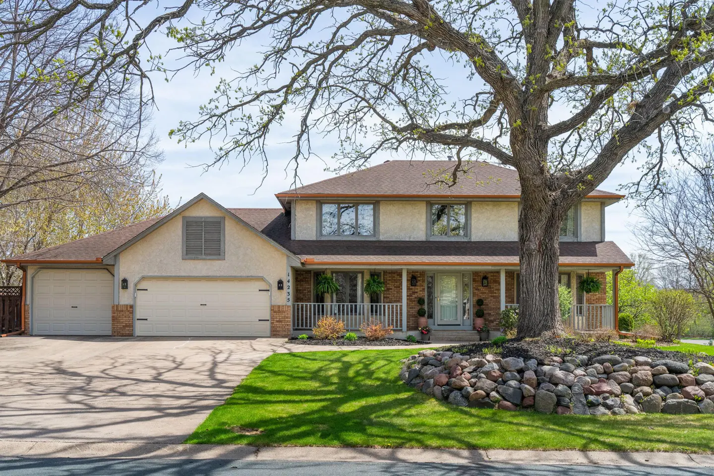 Two-story beige house with a brown roof and a three-car garage. A large tree stands in the front yard.