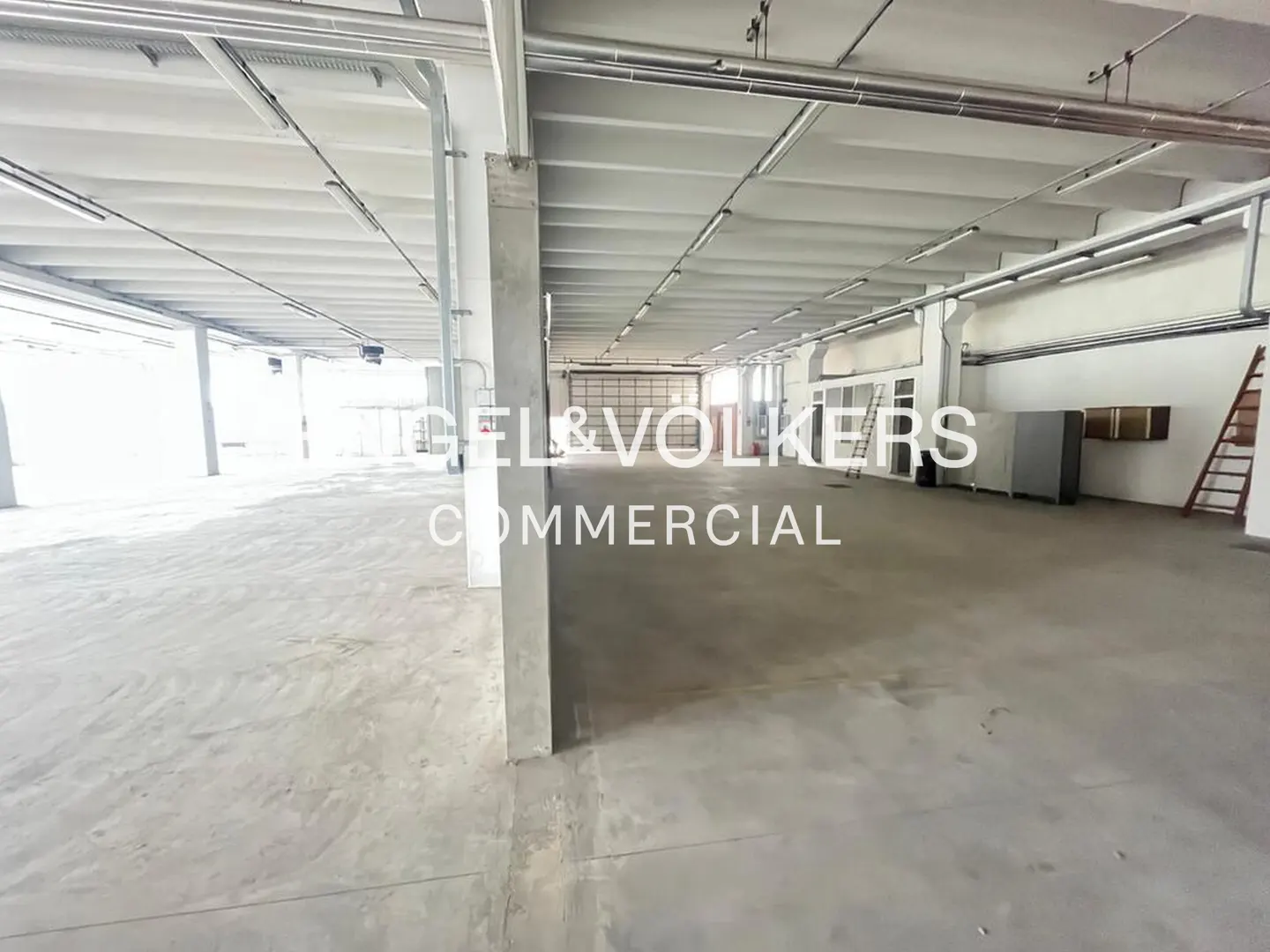 A large, empty commercial space with concrete floors and white walls. Overhead pipes and lighting fixtures are visible.