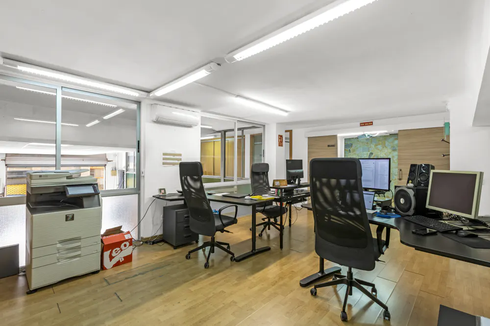 Bright office space with desks, black chairs, computers, and a printer. The floor is light wood, and there are large windows.