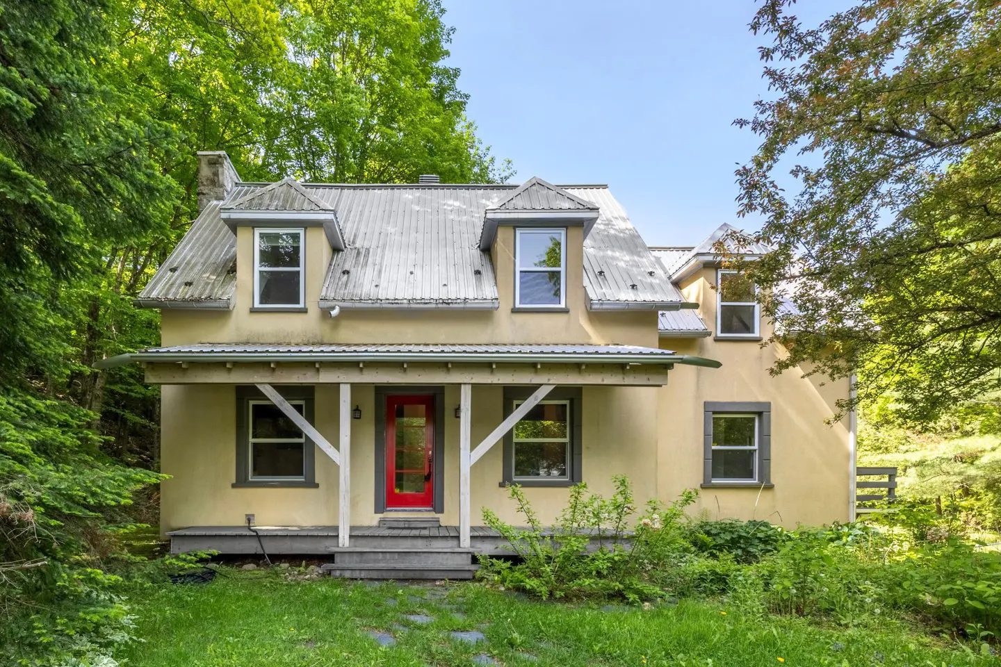 A two-story yellow cottage with a metal roof, a covered front porch, and a bright red front door, surrounded by lush trees.
