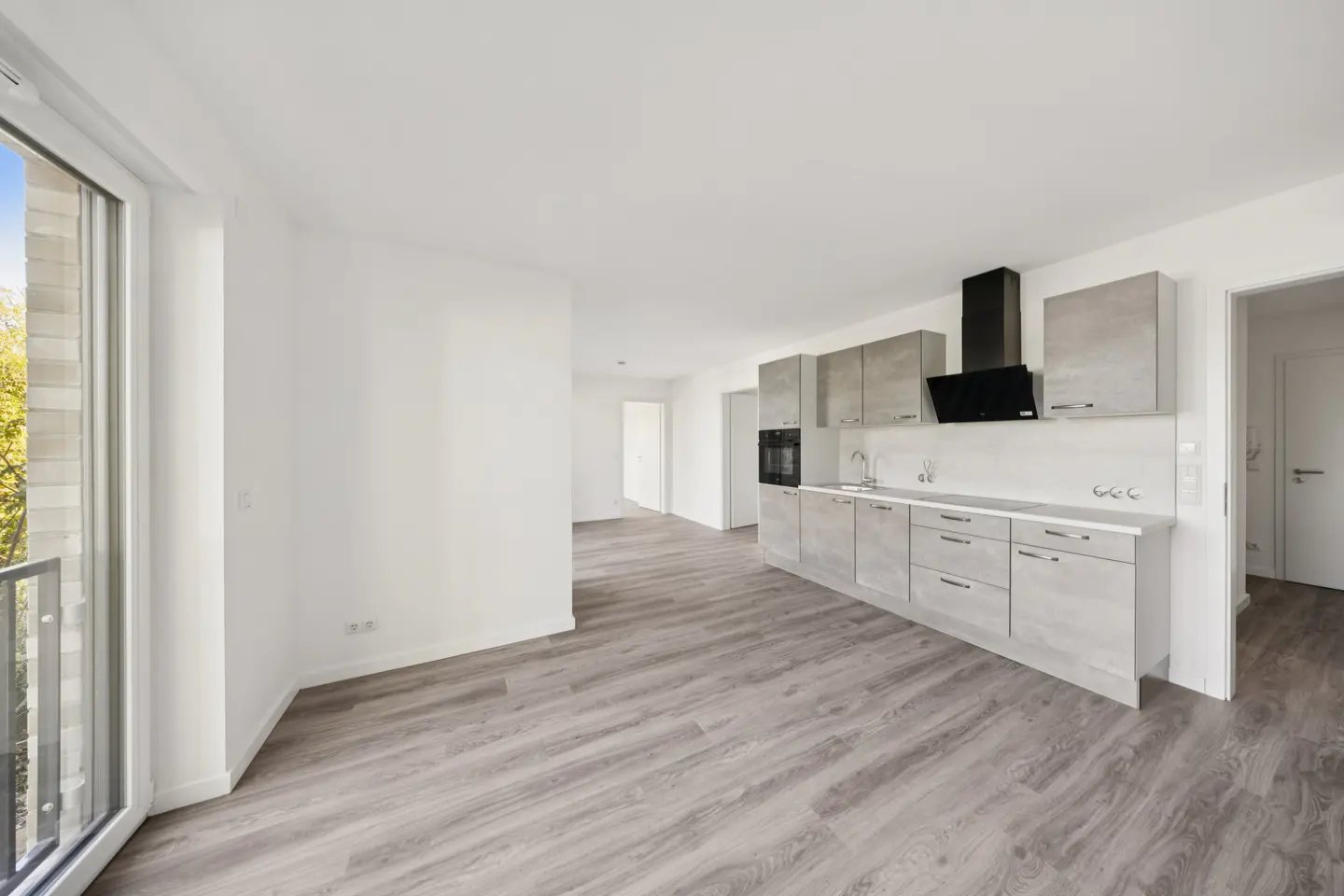 Bright, empty apartment with gray wood floors, white walls, and a modern gray kitchen with black range hood. Balcony door on the left.