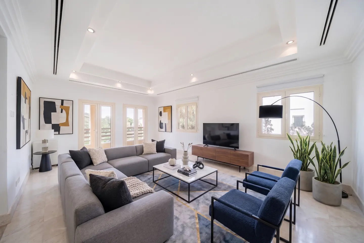 Modern living room with a grey sectional, two blue armchairs, a marble coffee table, and abstract wall art.