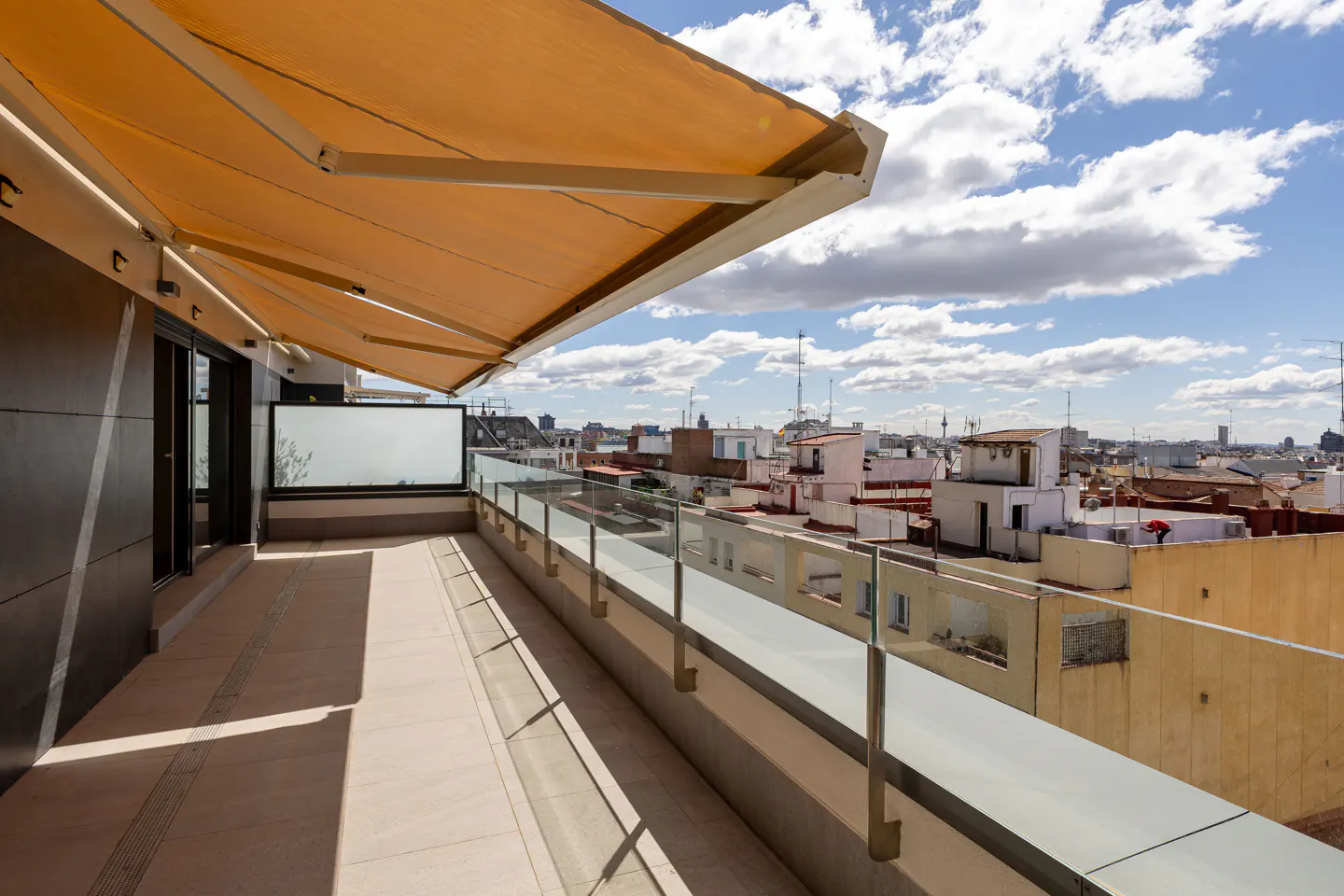 Balcony view with tan awning, glass railing, and city skyline under a blue sky with clouds.