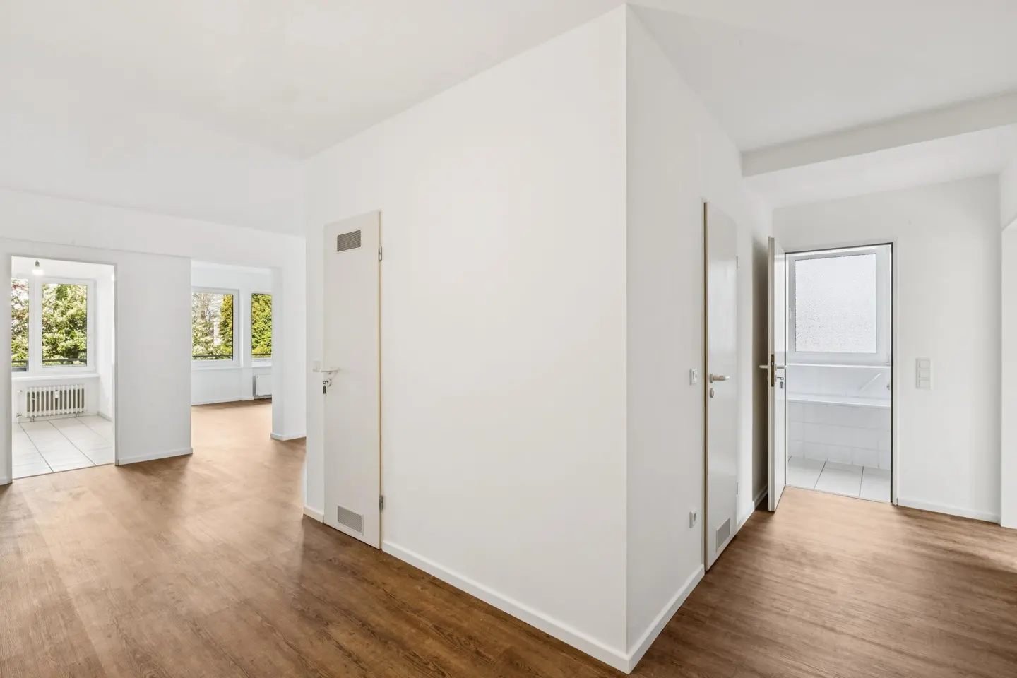 Bright, empty room with wood floors and white walls. Two doorways lead to rooms with windows. One door is open to a white bathroom.