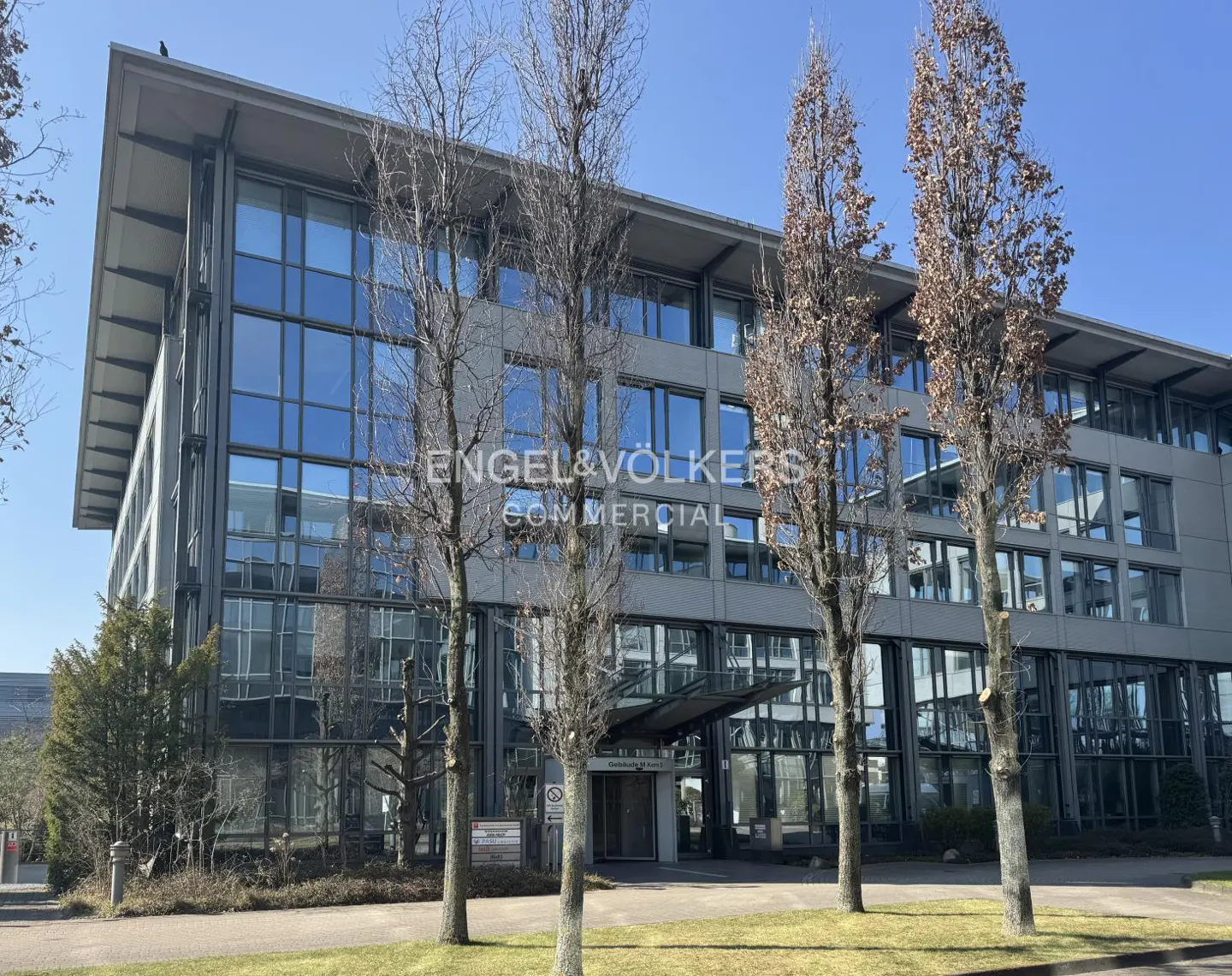 Exterior of a modern, multi-story commercial building with glass windows and Engel & Völkers signage. Trees line the front.