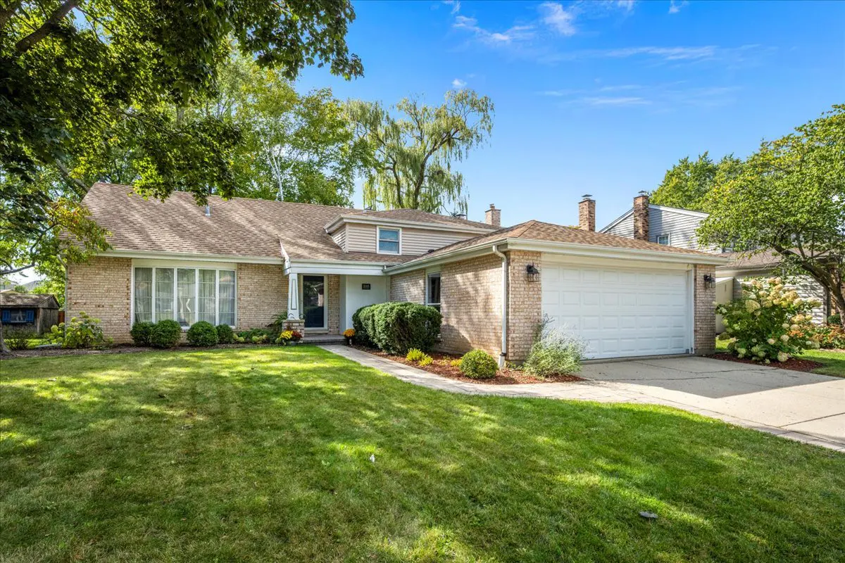 Single-story tan brick ranch home with a white garage, large front lawn, and mature trees under a bright blue sky.
