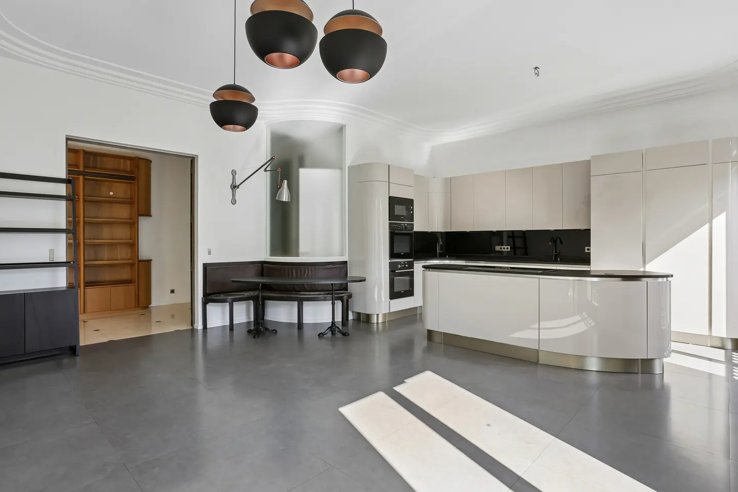 A modern kitchen with gray floors, white cabinets, and black countertops. A dark brown booth seating area is next to the kitchen. Three black pendant lights hang from the ceiling.
