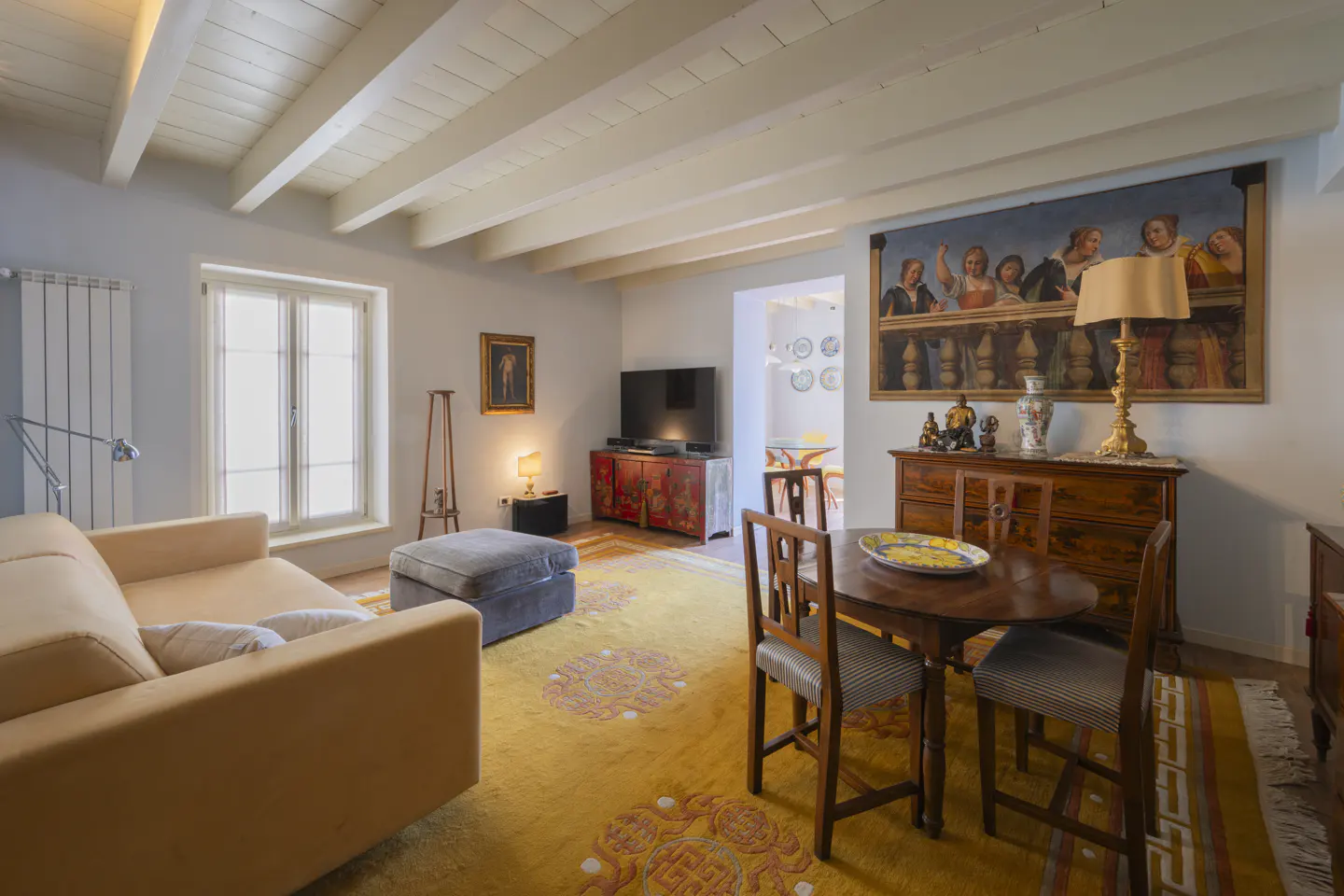 Living room with a beige sofa, wooden table and chairs, yellow rug, and a painting on the wall.