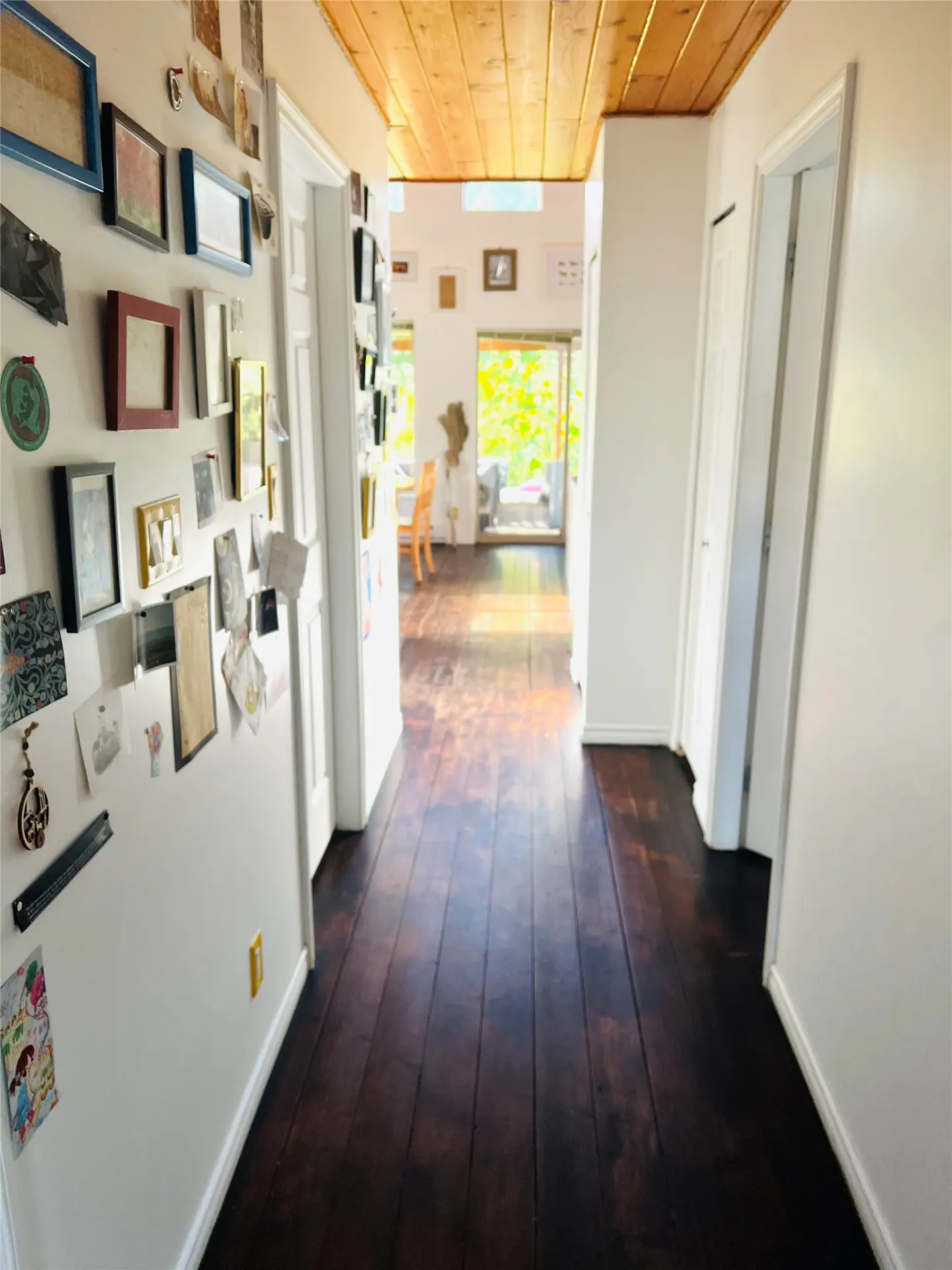 Hallway view with dark wood floors, white walls, and a wood plank ceiling. A wall covered in framed photos and art is on the left.