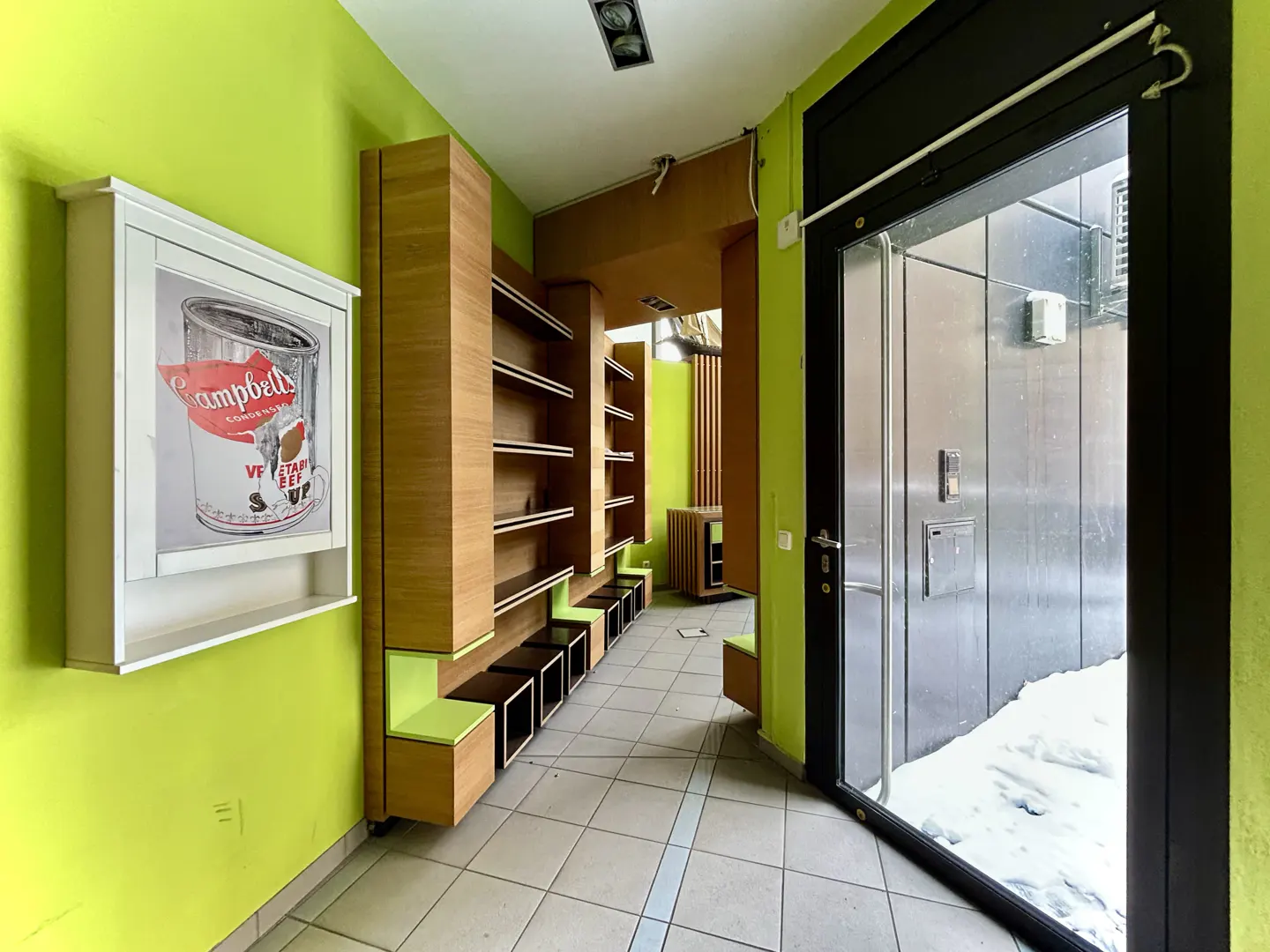 Interior view of a commercial space with lime green walls, wooden shelving, and a black-framed glass door showing snow outside.