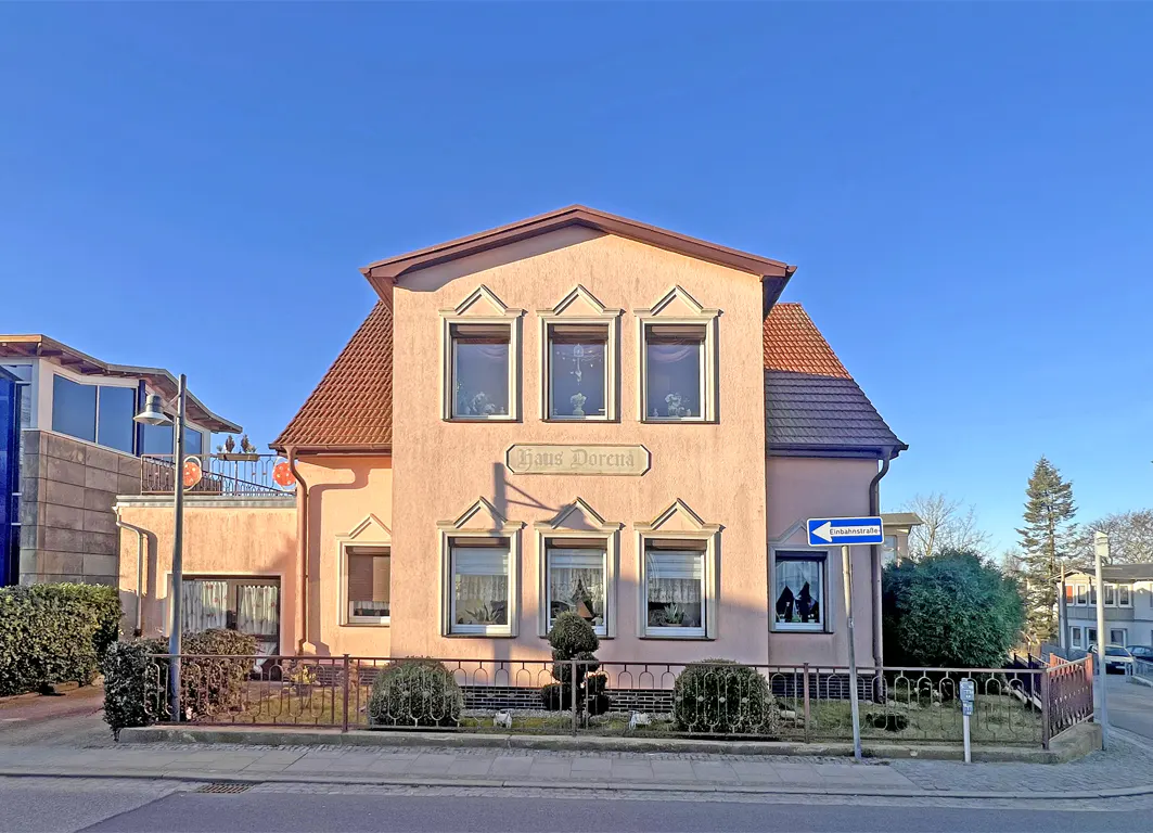 A two-story peach house with a red roof, named "Haus Dorena," sits behind a black metal fence under a clear blue sky.