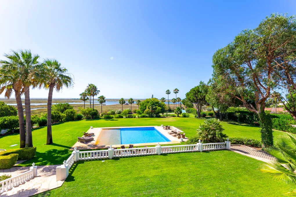 Aerial view of a blue swimming pool surrounded by green grass, palm trees, and a white fence under a clear blue sky.