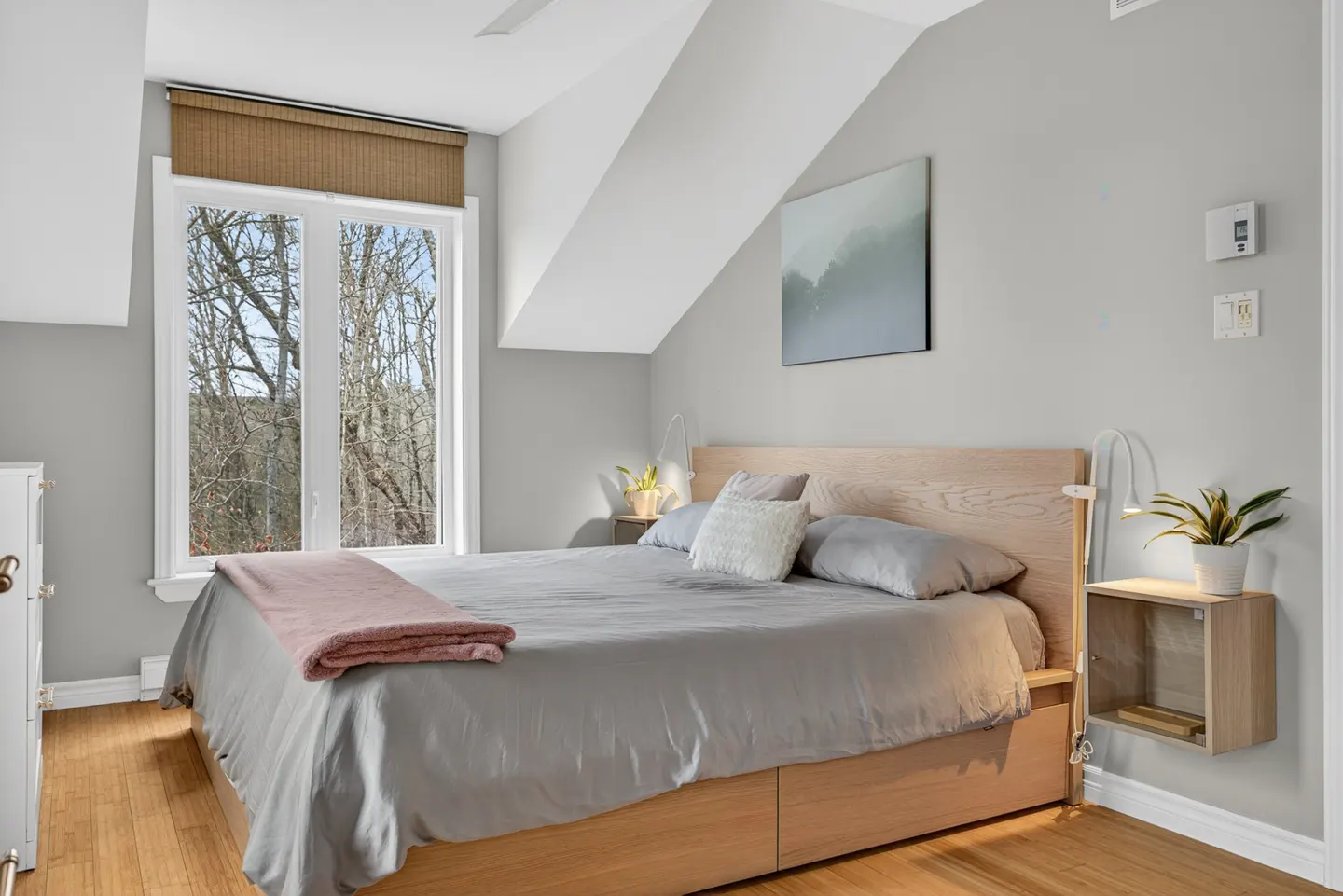 Bedroom with gray walls, a light wood bed frame, gray bedding, and a pink throw blanket. A window with a brown shade is on the left.