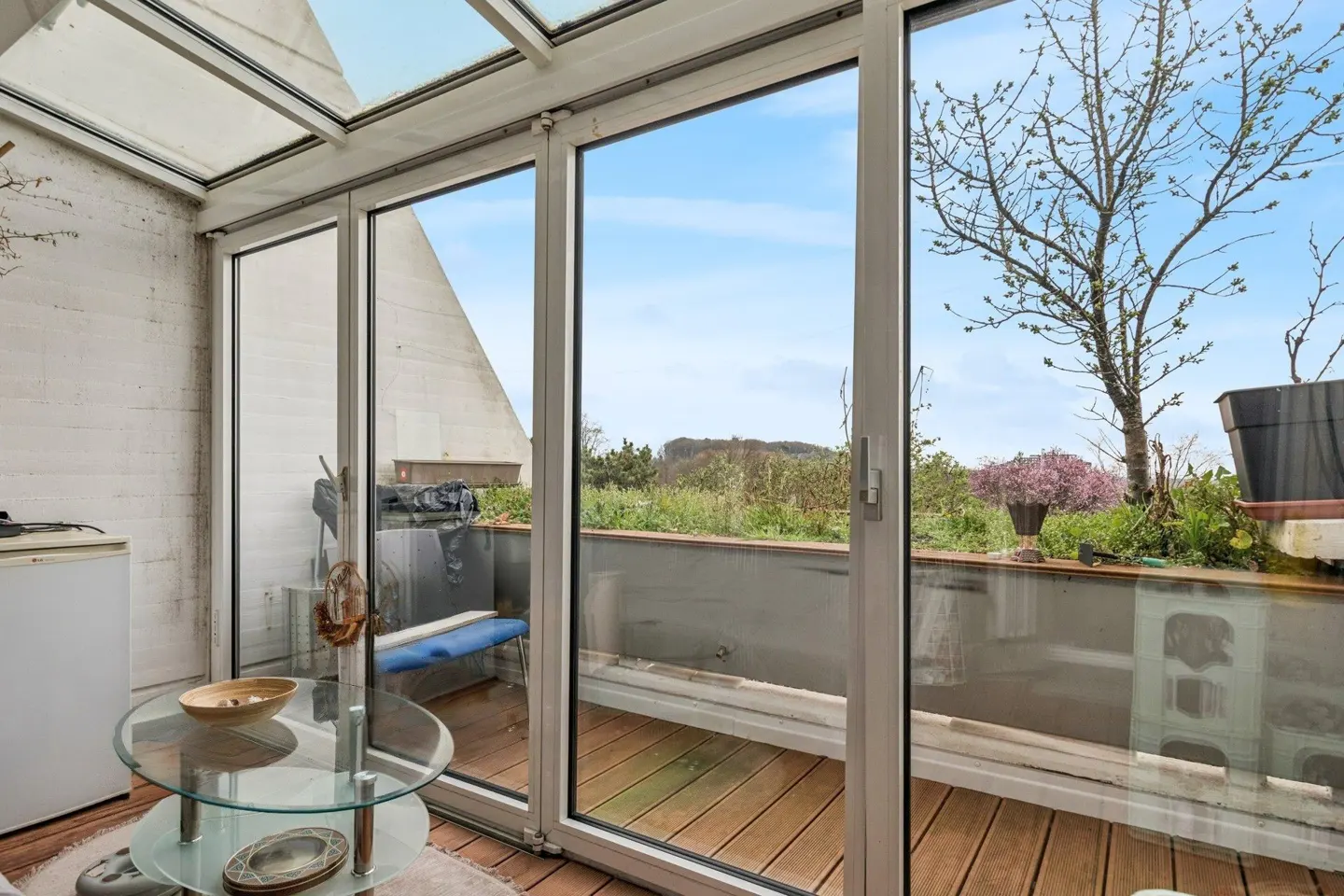 Sunroom with glass roof and sliding doors to a wood deck. A glass table sits in the room, with a view of trees and sky outside.