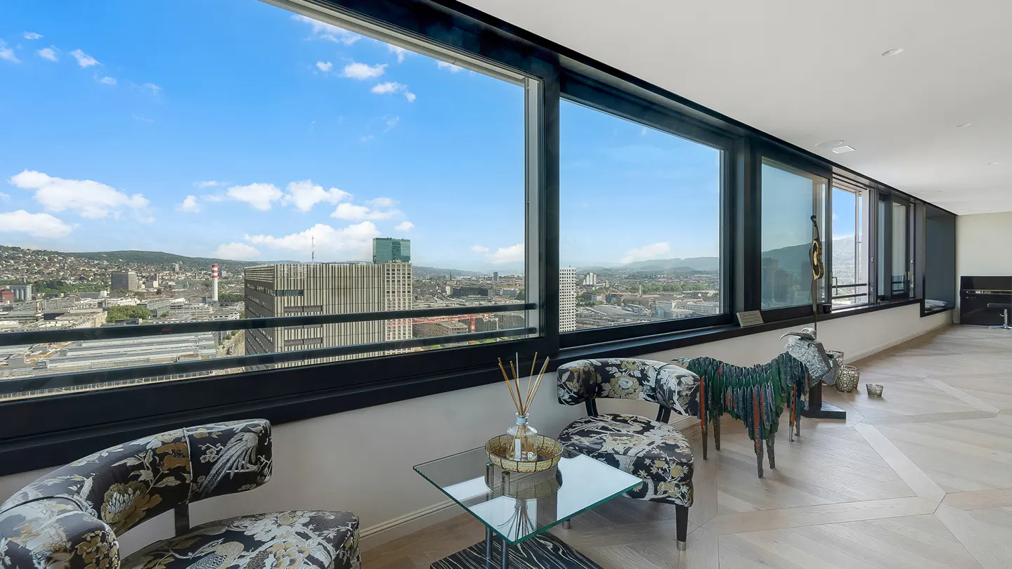 A high-rise apartment interior with floral chairs, a glass table, and a panoramic city view through large windows.