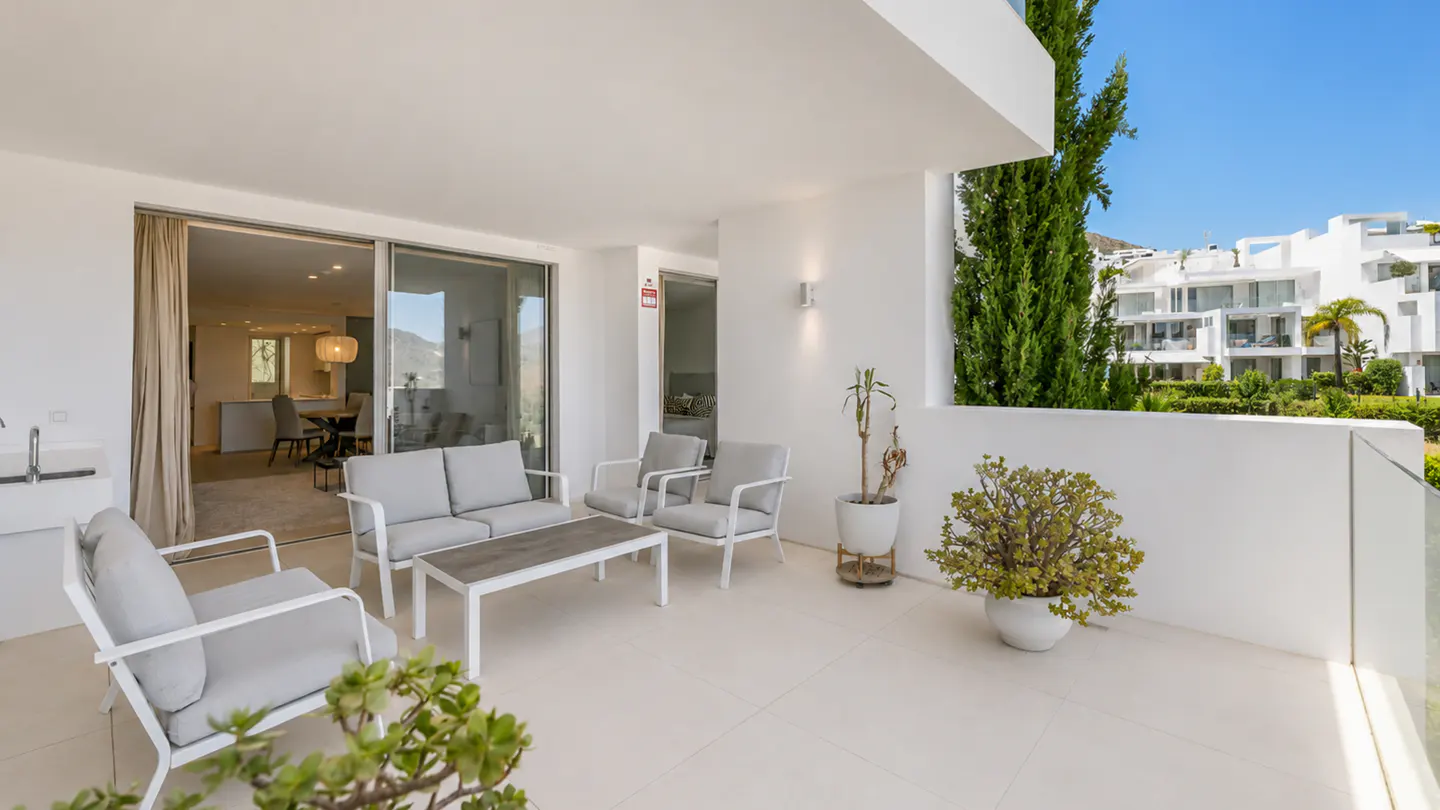 Modern white terrace with grey outdoor furniture, potted plants, and a view of a bright residential complex.