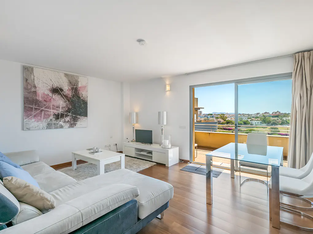 Bright living room with a gray and white sofa, a glass table, and a balcony overlooking a cityscape.