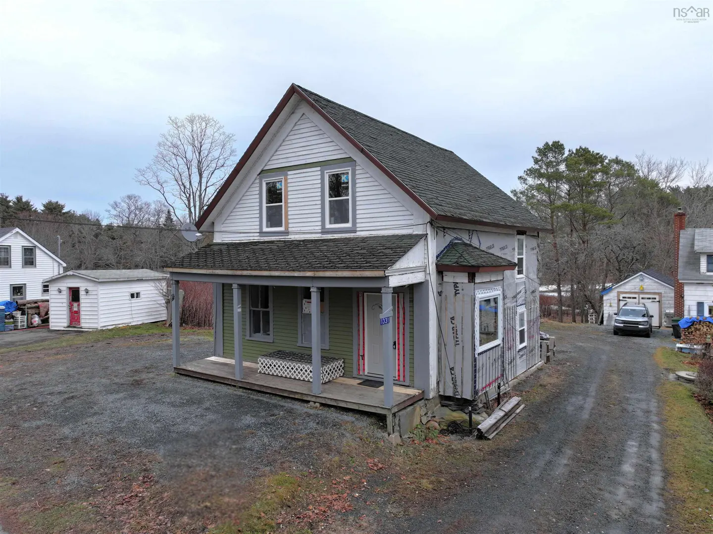 Two-story house with a covered porch, gray and green siding, and a dark roof. Some siding is unfinished. Gravel driveway.