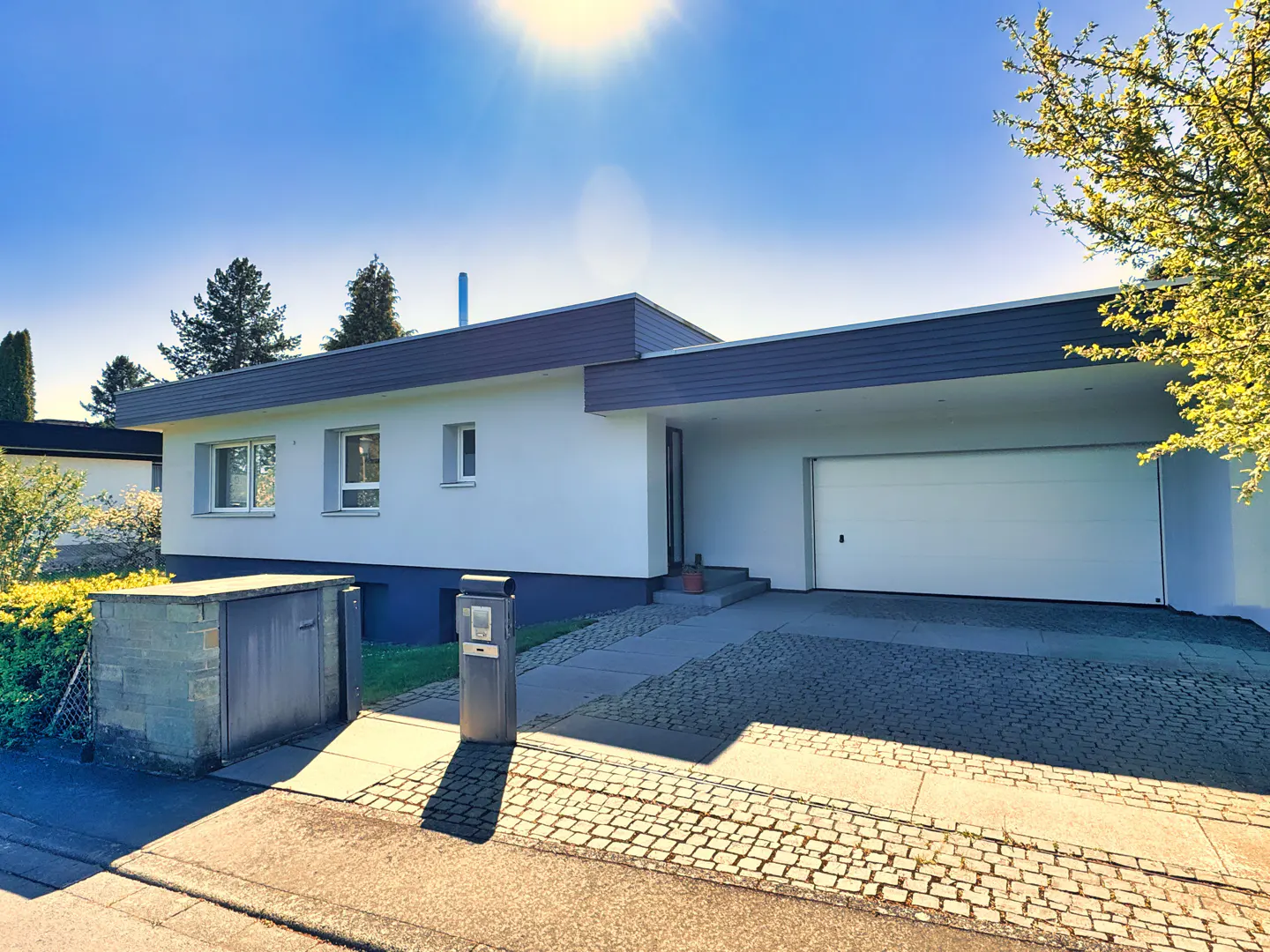Exterior of a modern, single-story white house with a gray roof and a white garage door on a sunny day.