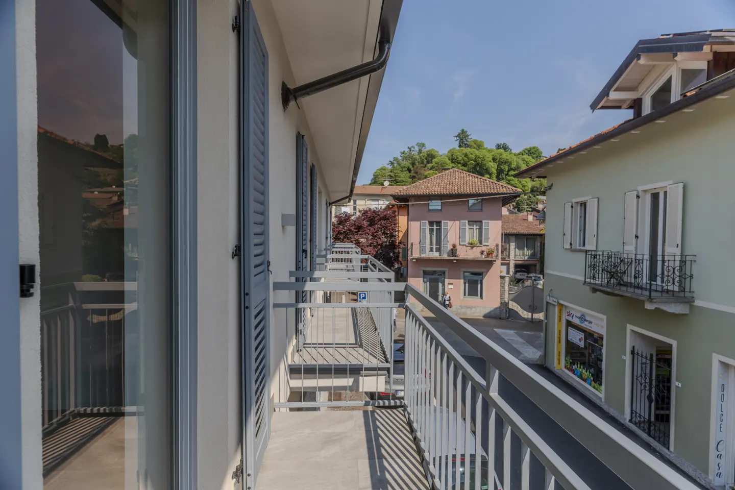 View from a balcony with gray railings and blue shutters, overlooking buildings and a street on a sunny day.