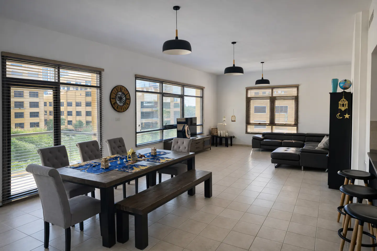 Bright, open-plan living and dining area with large windows, a dark wood dining table with gray chairs, and a black leather sofa.