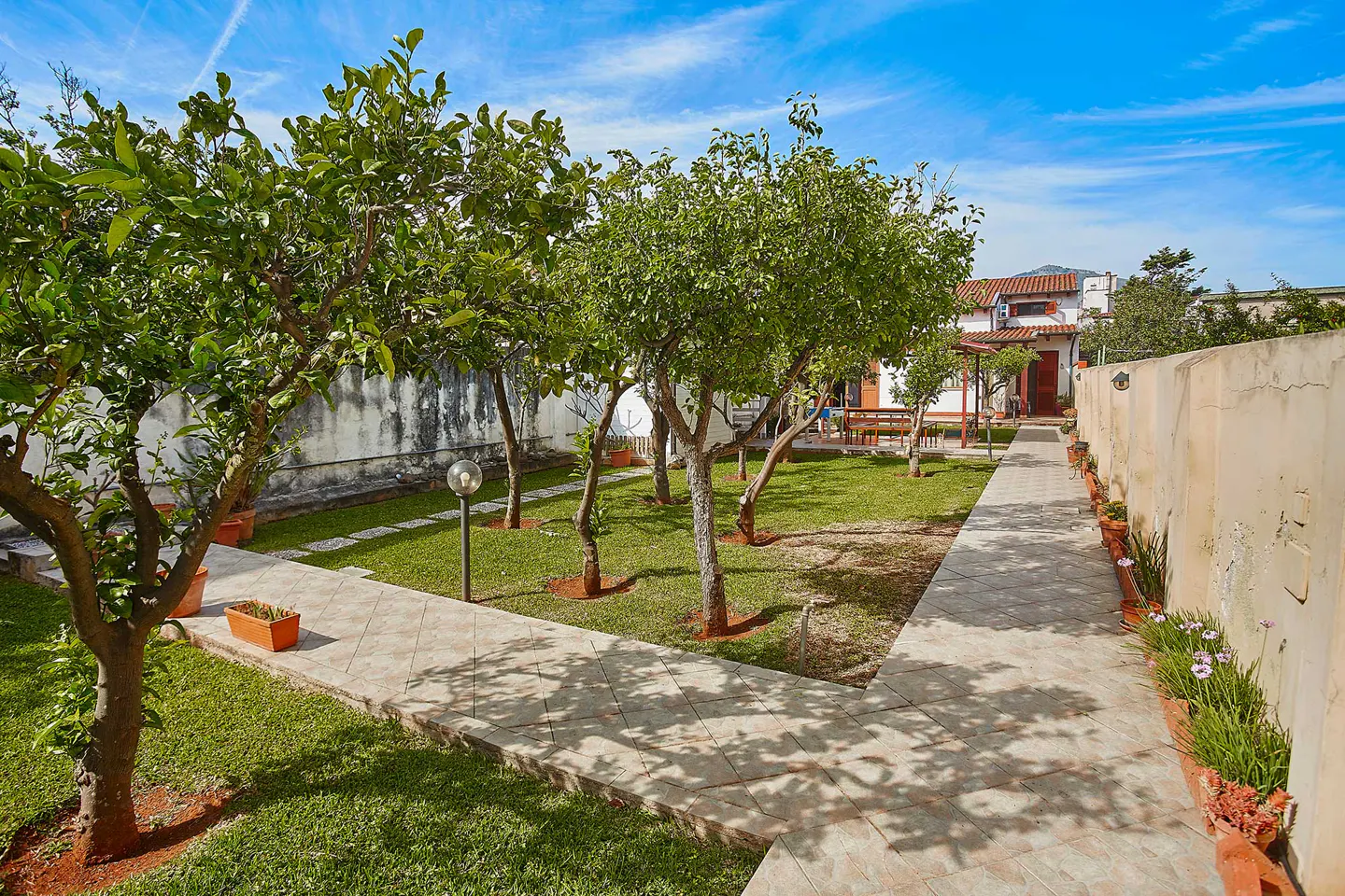 A backyard with a tiled path, green grass, and trees leading to a house under a blue sky.