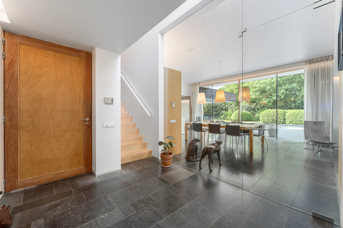 Modern foyer with a wooden door, light wood staircase, and a glass-walled dining room with a table and garden view.
