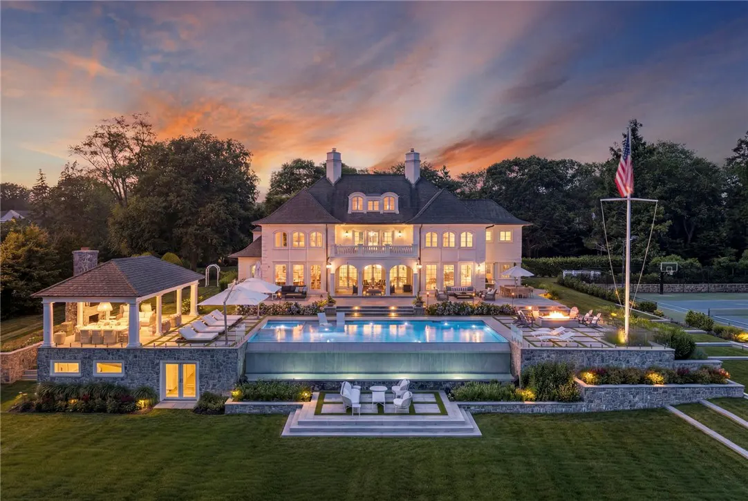 A large, cream-colored house with a pool and a gazebo at sunset. An American flag flies in the background.