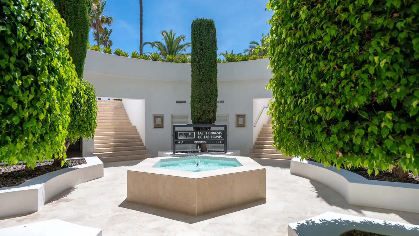 A hexagonal fountain sits in a courtyard with white walls, stairs, and lush green trees under a bright blue sky.