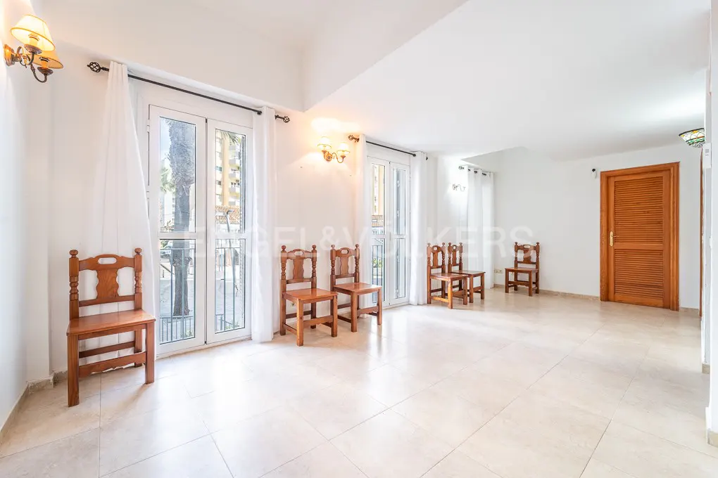 Bright, empty room with white walls, tile floor, and wooden chairs. French doors with white curtains lead to a balcony. A wooden door is on the right.