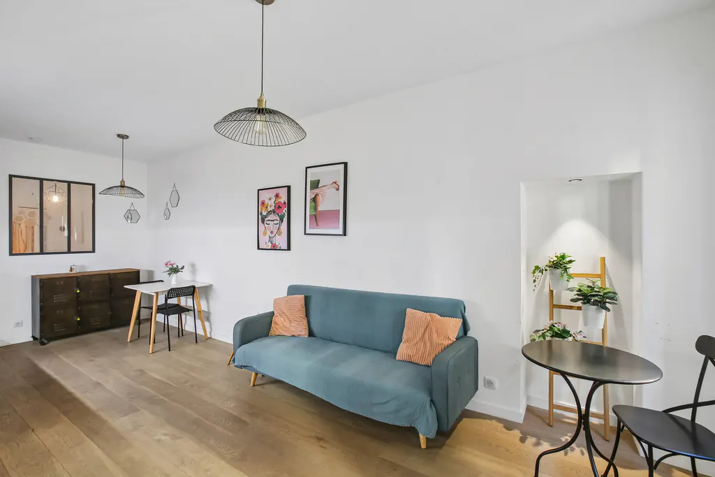 Bright living room with wood floors, white walls, and modern decor. A blue sofa with orange pillows sits near a black table and chairs. Artwork adorns the walls.