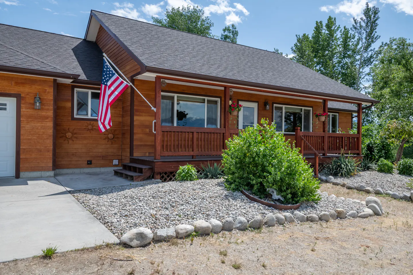 Exterior view of a one-story wood house with a porch, American flag, gray roof, and rock landscaping.