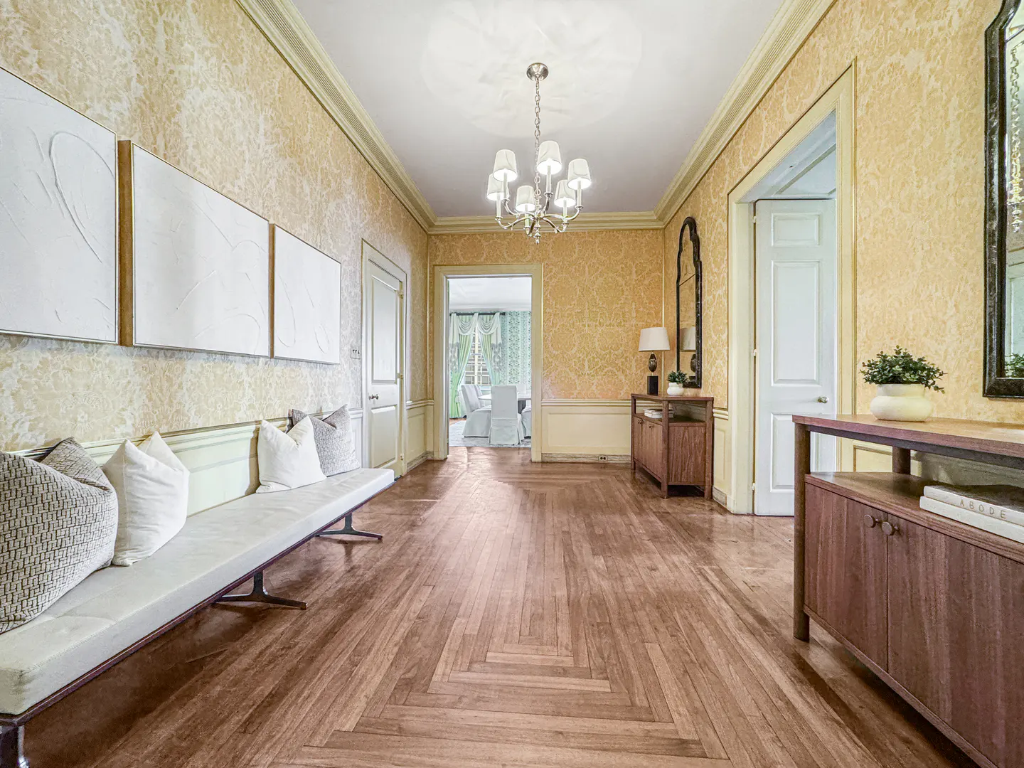 Hallway with parquet floors, patterned wallpaper, and white trim. A bench with pillows sits under three white paintings. A chandelier hangs from the ceiling.