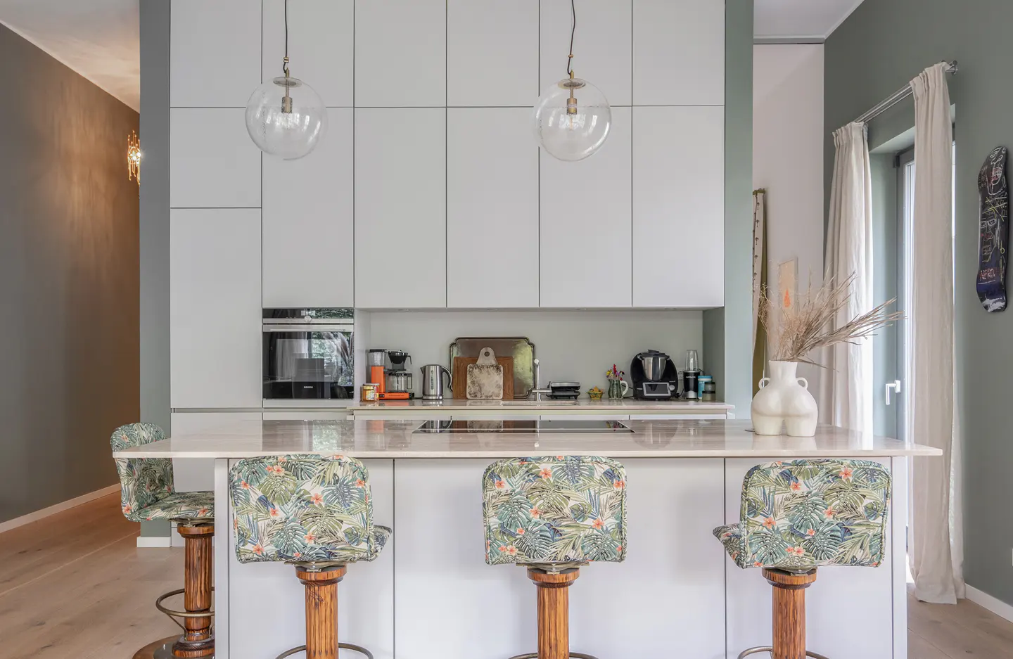 Bright kitchen with white cabinets, marble countertop island, and three floral-patterned bar stools. Two globe pendant lights hang above.