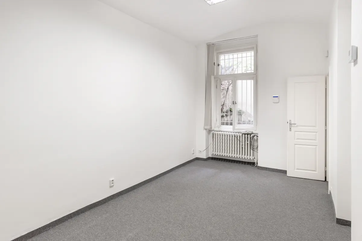 Empty room with white walls, gray carpet, and a window with a radiator. A white door is on the right.