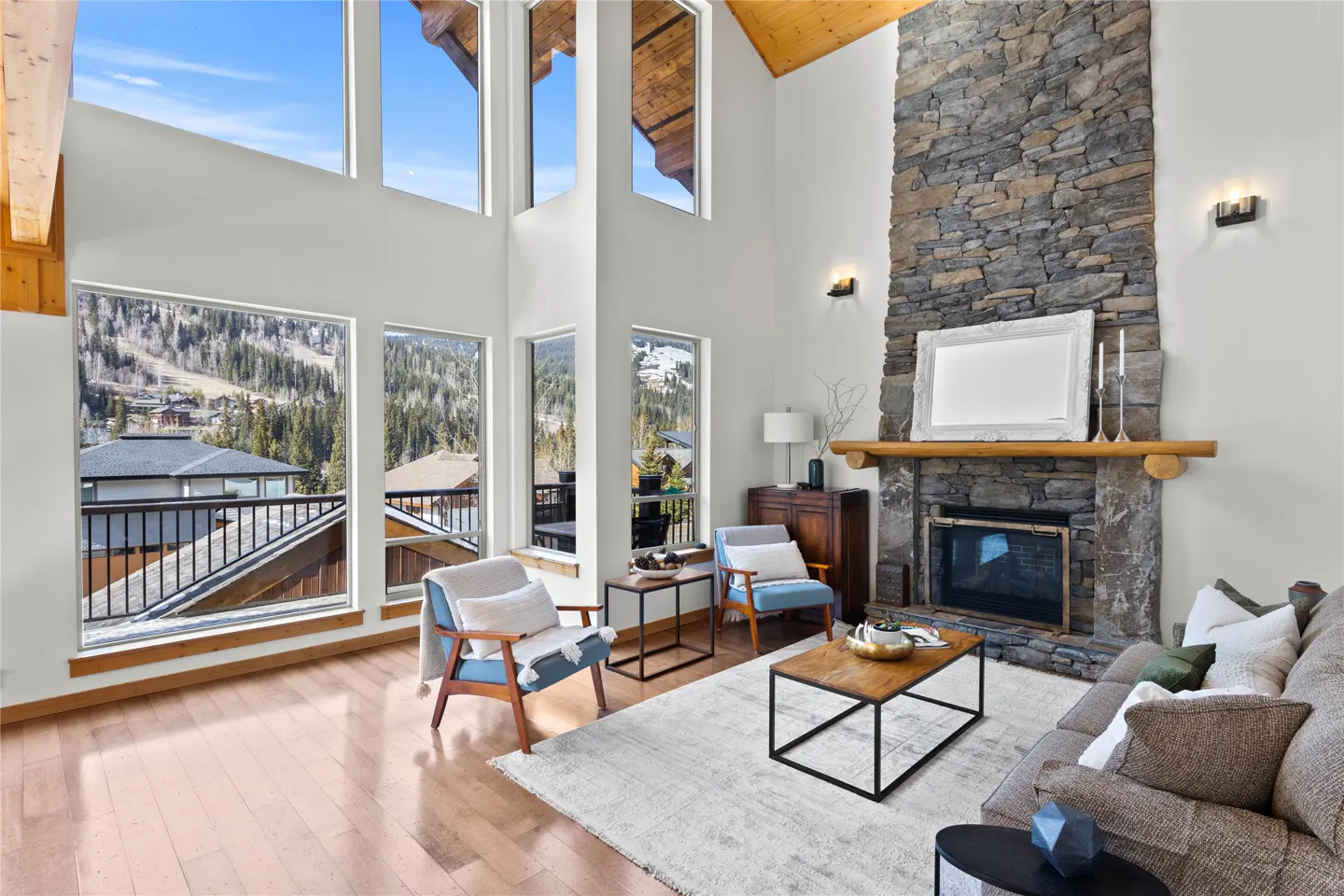 Bright living room with stone fireplace, wood floors, and large windows with mountain views. Blue chairs and a gray sofa furnish the space.