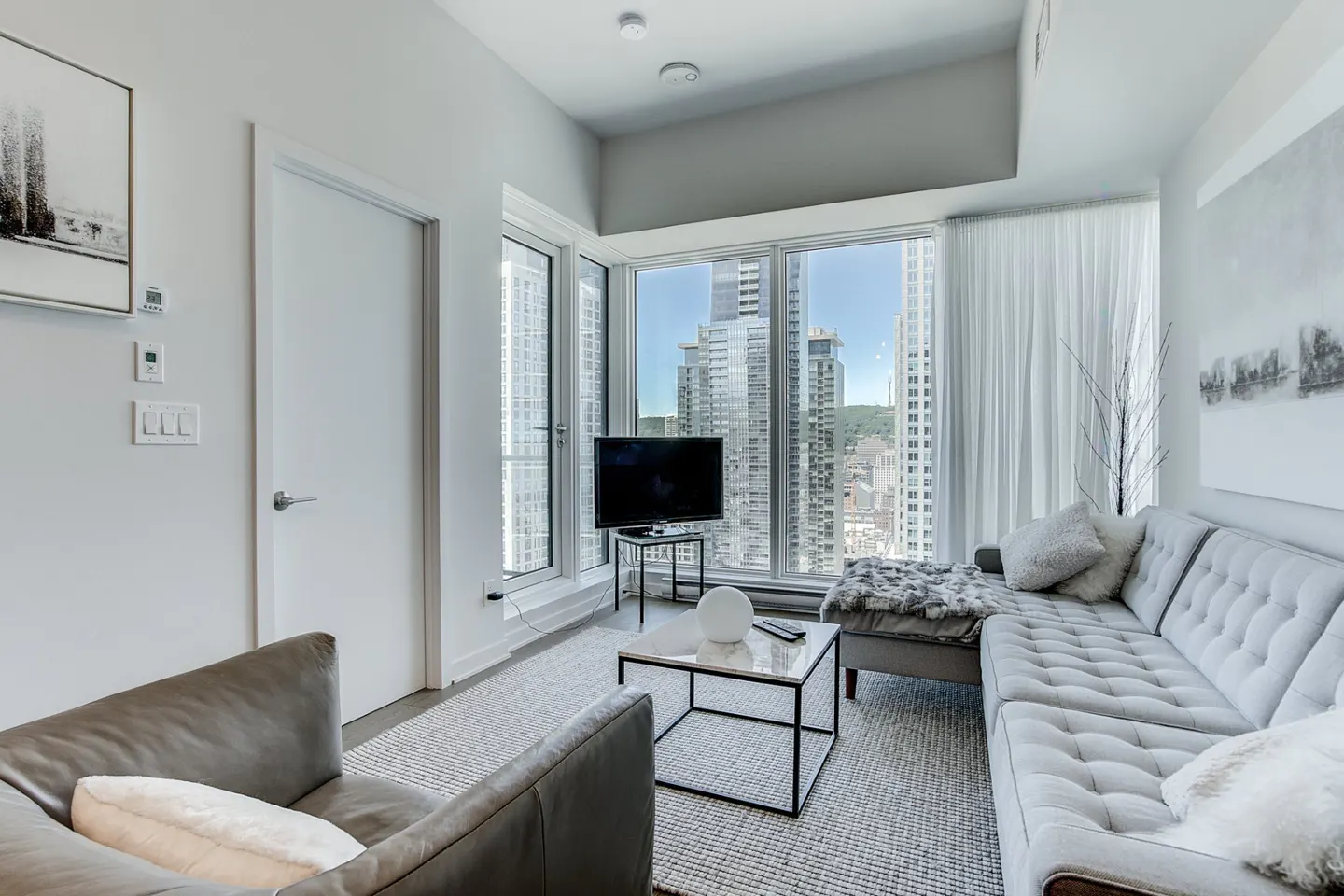 Living room with gray sofa, leather chair, and city view. A TV sits by the window, and a marble table is on a gray rug.