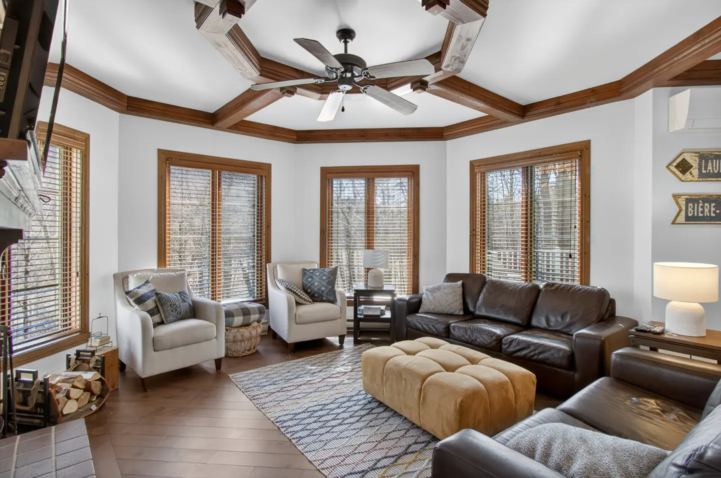 Living room with brown leather sofas, beige chairs, and wood-framed windows. A ceiling fan hangs from a wood-beamed ceiling.